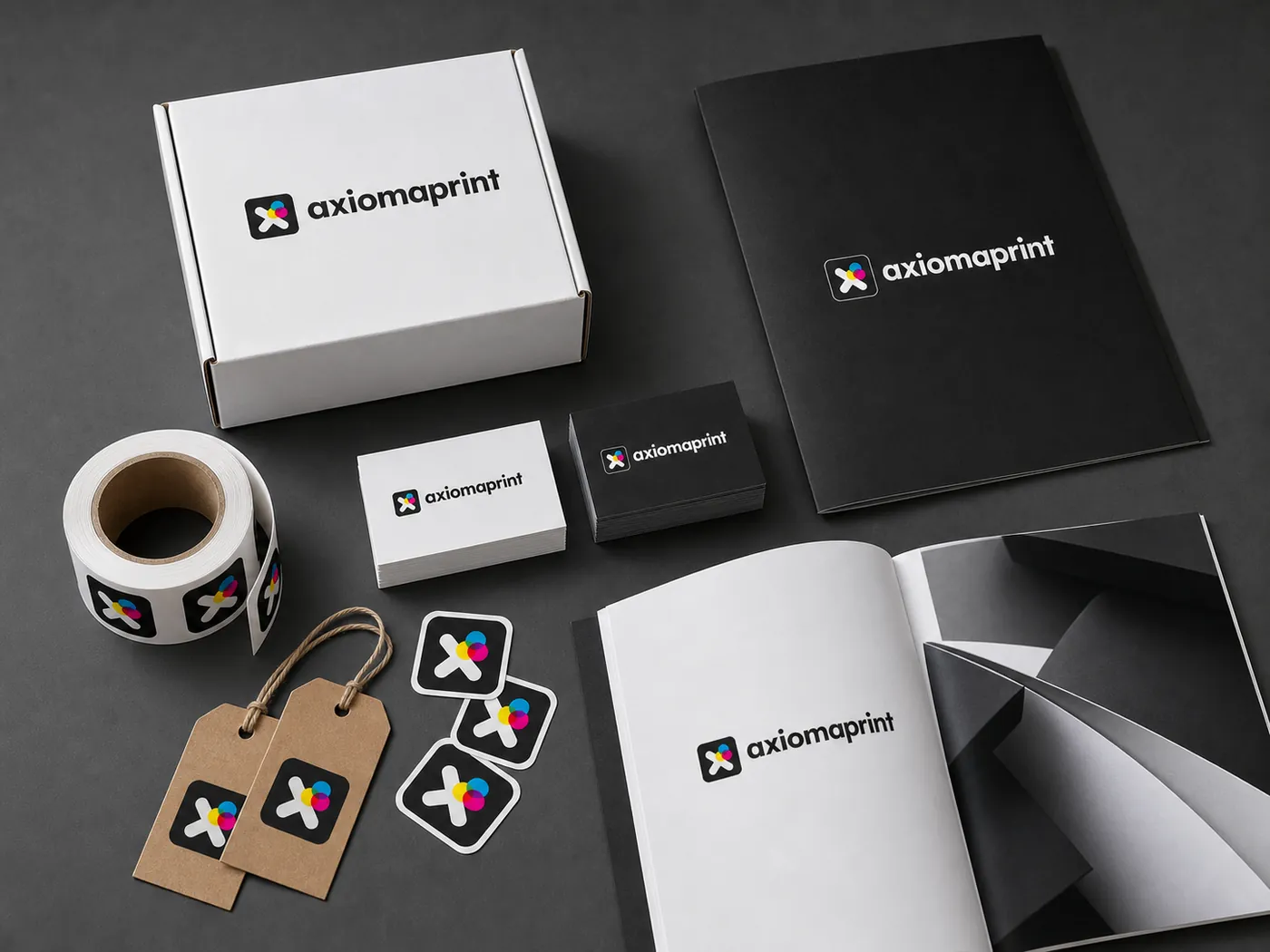 Caja blanca, carpeta negra, rollo de pegatinas, etiquetas colgantes de cartón, dos tacos de tarjetas y un catálogo abierto sobre fondo gris