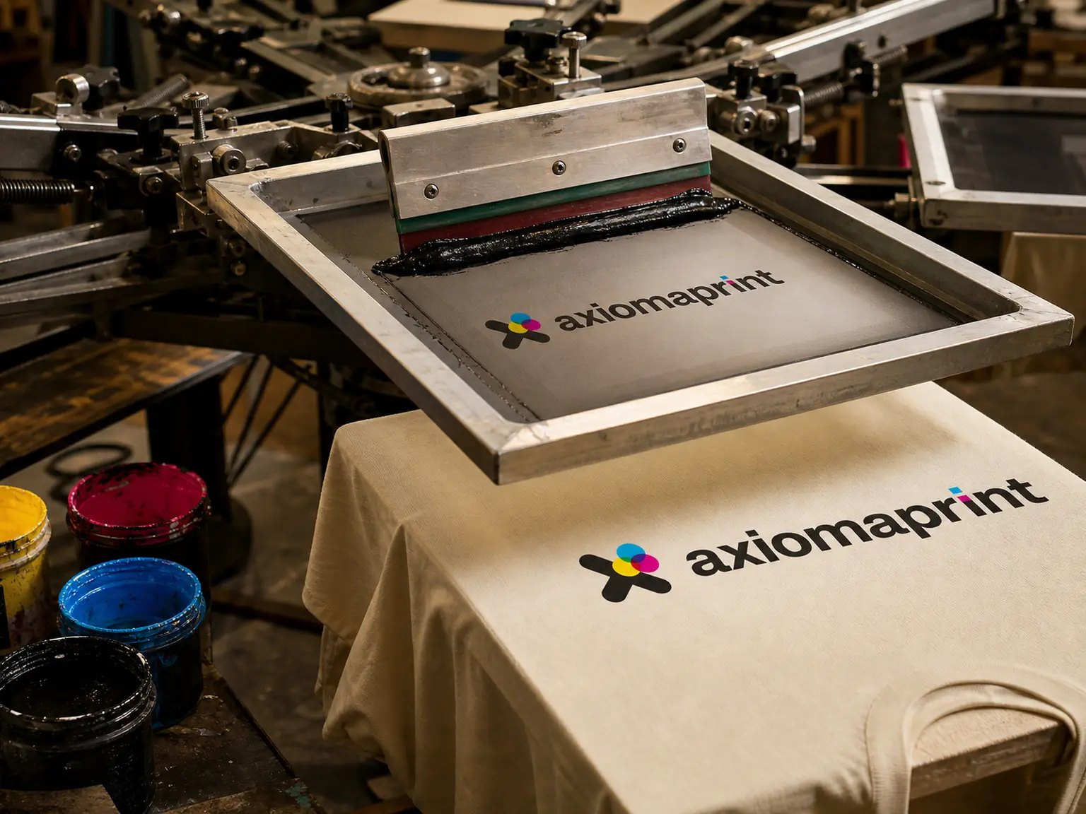 Pantalla de serigrafía con rasqueta y tinta negra sobre una camiseta cruda recién impresa, junto a botes de tinta amarilla, roja y azul